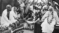 At a formal function, an aged bald man and an old women are humbly dressed and seated side-by-side with legs folded on a rug-strewn dais at right; the man looks at a bearded, robed, and garlanded old man seated on another dais at left, who is reading from a sheet of paper held in his left hand. In the foreground, various dishes and ceremonial objects are arrayed; in the background, a half-dozen dignitaries and dozens of ordinary people observe.